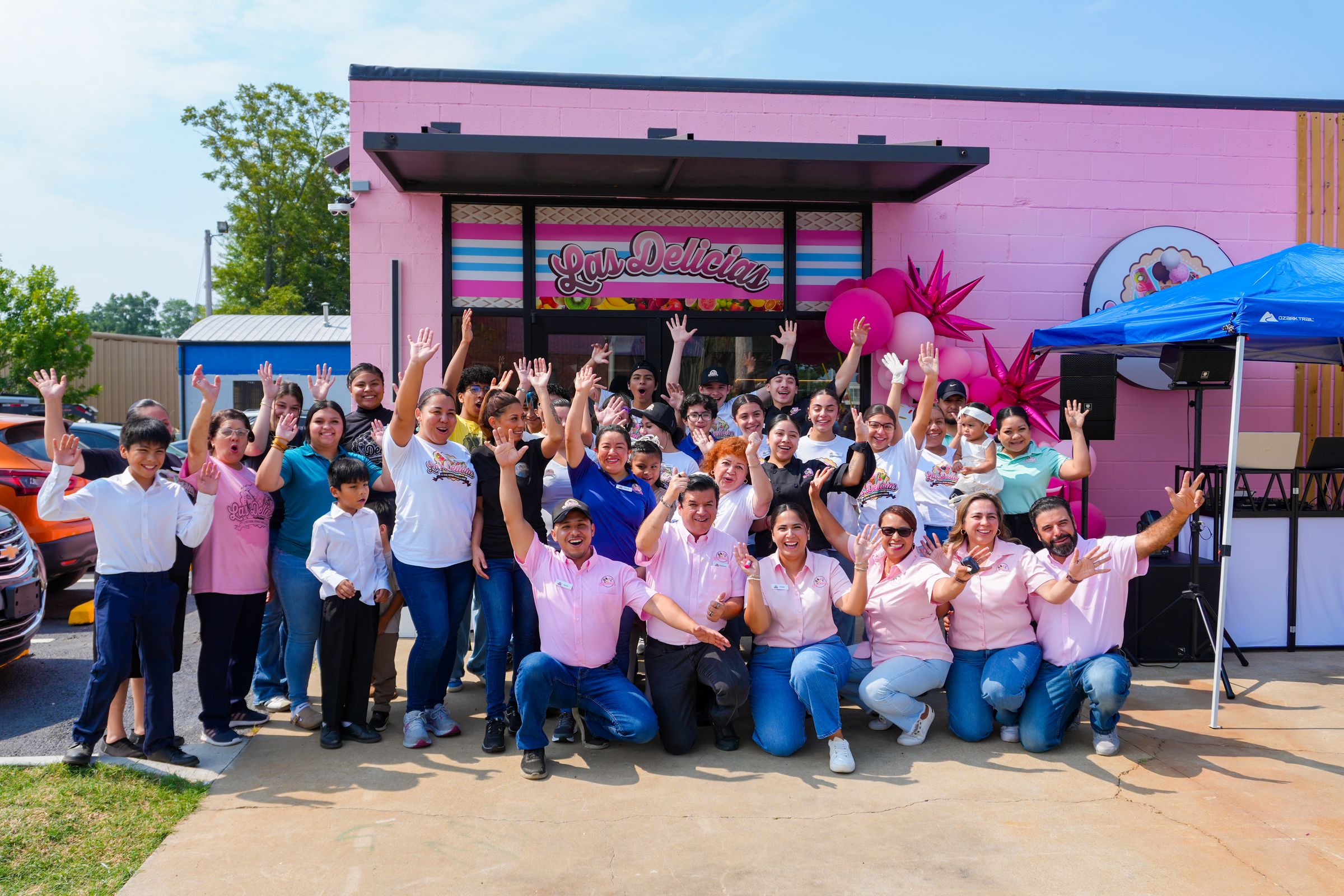 Las Delicias employees, families, and community members celebrating with their hands raised outside the pink storefront