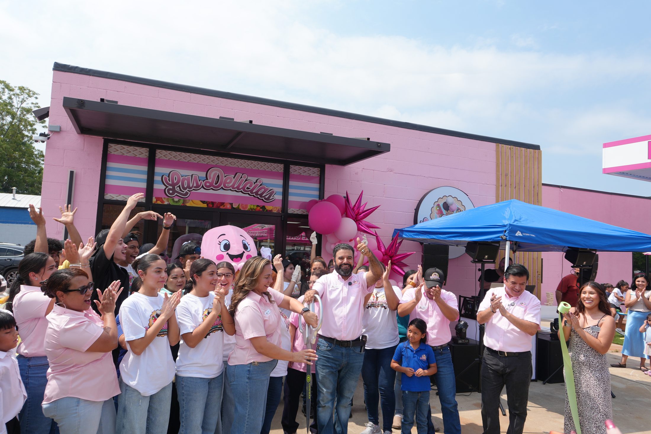 La familia y el equipo cortando el listón afuera de la tienda rosa de la calle Harkrider, con la mascota rosa de helado detrás