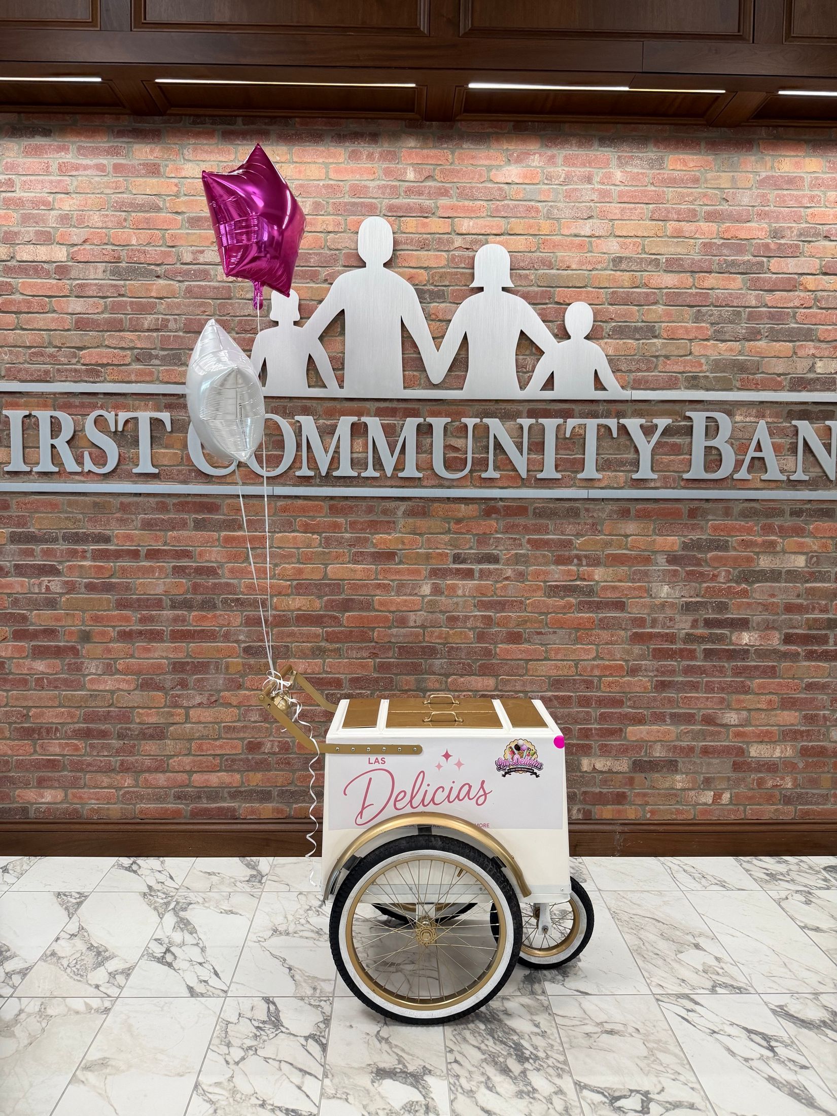 The white and gold Las Delicias push cart parked outside a brick building with balloons tied to the handle